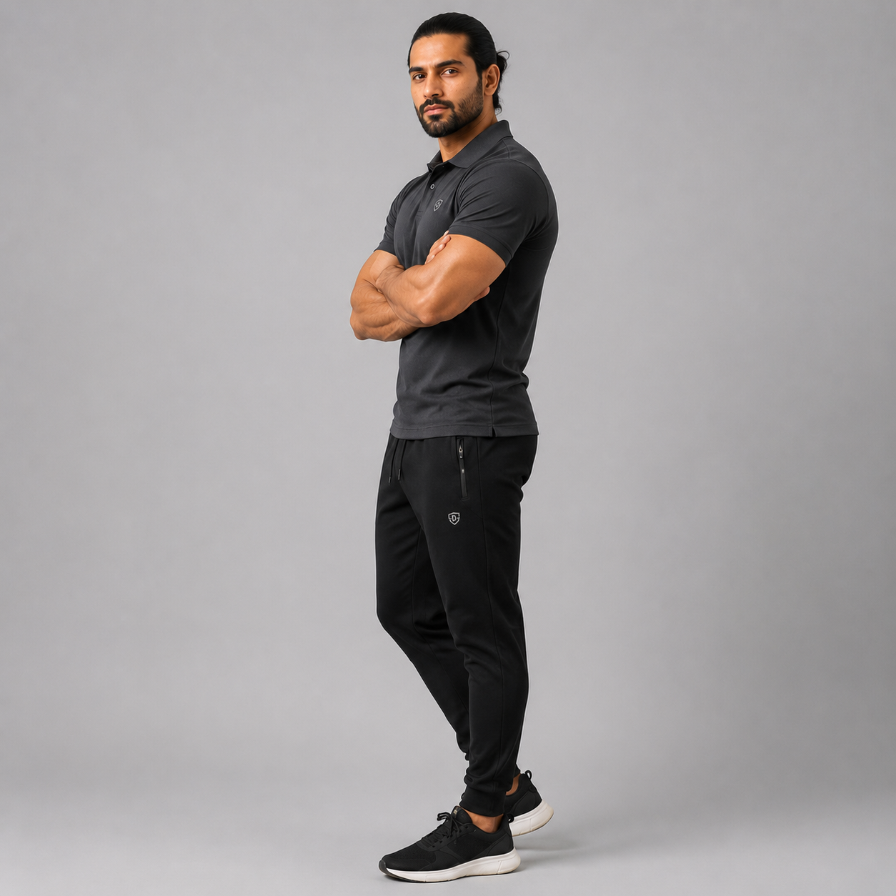 A man modeling a dark gray short-sleeve polo shirt and black jogger pants with black sneakers, standing against a solid gray studio background.
