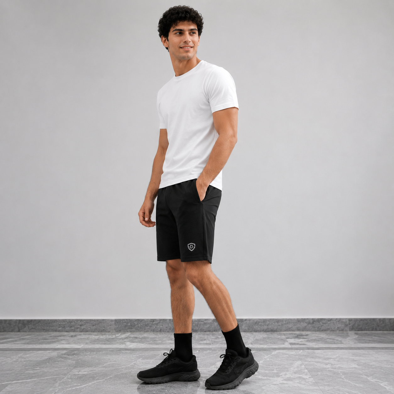 A man wearing a white crew-neck t-shirt, black athletic shorts with a small shield logo on the left leg, black socks, and black sneakers, standing against a light gray wall and tiled floor.
