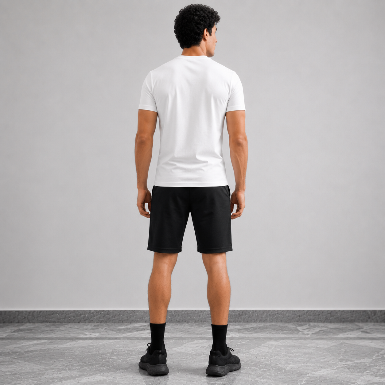 A man viewed from behind wearing a white short-sleeve t-shirt, black athletic shorts, black socks, and black sneakers, standing against a light gray studio wall.