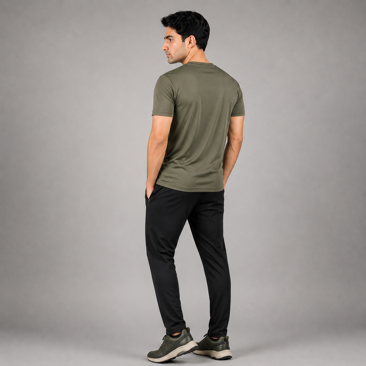 A man wearing an olive green short-sleeve t-shirt and black pants, shown from a rear three-quarter view against a solid gray studio background.