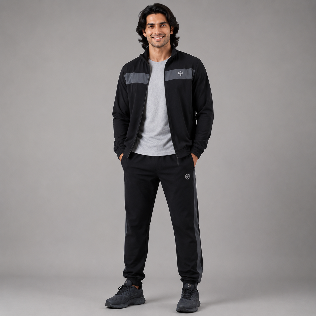 A man modeling a black athletic tracksuit set, featuring a zip-up jacket with gray chest panels and matching black sweatpants, styled over a white t-shirt and dark sneakers against a solid gray studio background.