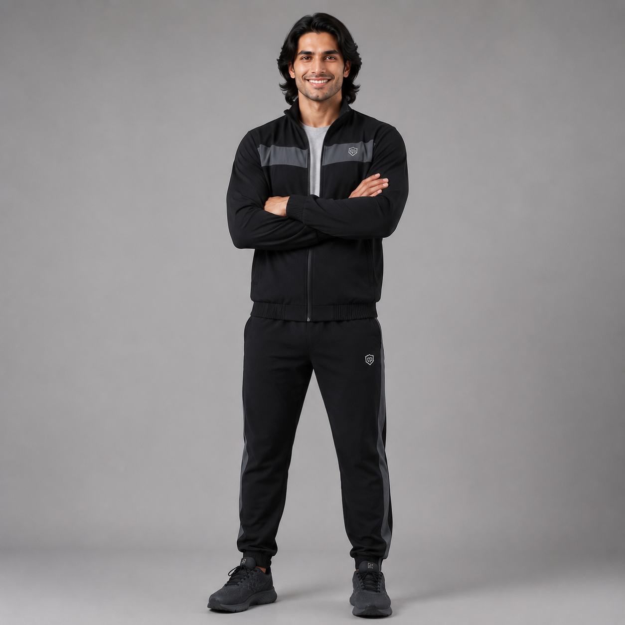 A man modeling a black athletic tracksuit with gray shoulder panels and a small shield logo on the chest and thigh, standing against a neutral gray studio background.