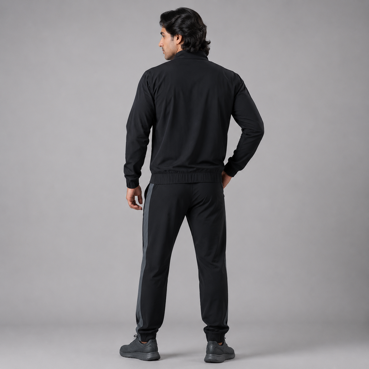 A man wearing a black tracksuit, viewed from behind, standing against a solid gray studio background.