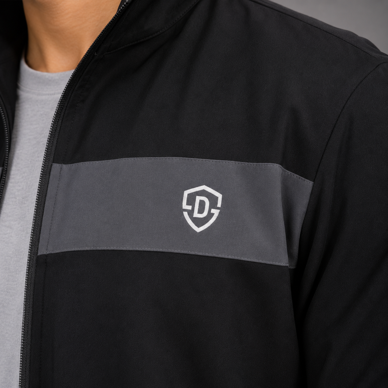 Close-up of a black zip-up jacket worn by a model, featuring a horizontal gray chest stripe with a white shield logo containing the letter D.