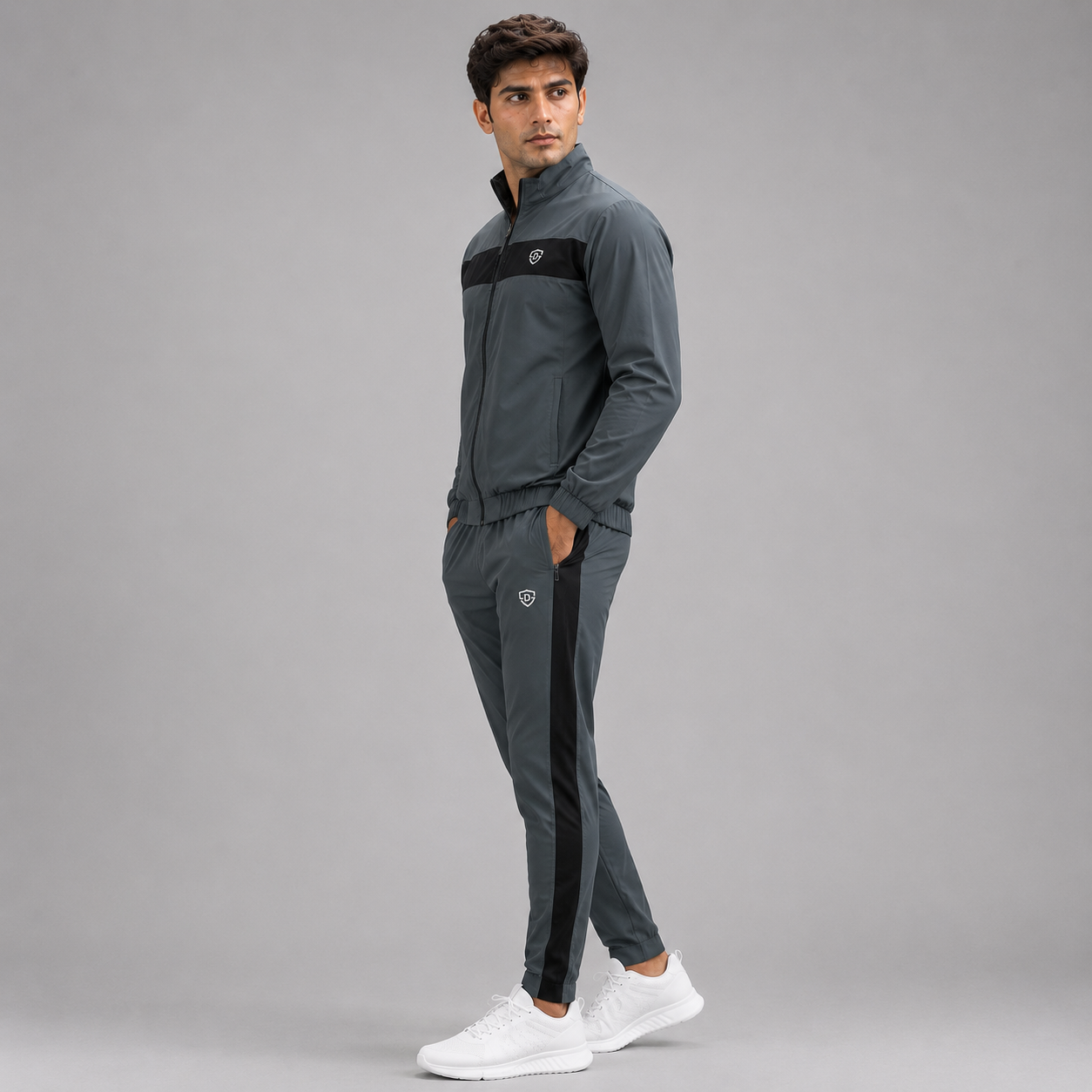 A man modeling a dark gray athletic tracksuit with black stripe accents on the chest and down the sides of the pants, paired with white sneakers, against a solid gray studio background.