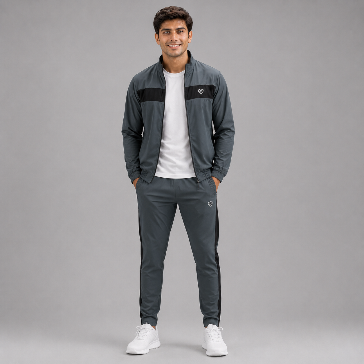 A man modeling a dark gray athletic tracksuit with black stripe details, a white t-shirt, and white sneakers against a solid gray studio background.