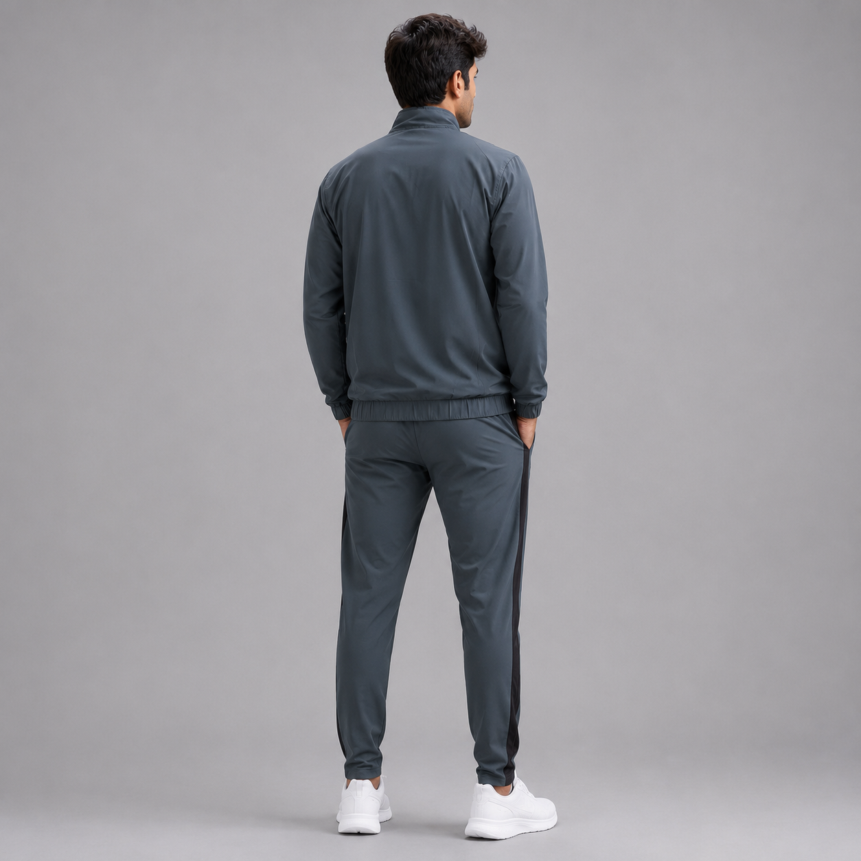 A man viewed from behind wearing a dark gray athletic tracksuit with a zip-up jacket and matching jogger pants, paired with white sneakers, against a solid gray studio background.