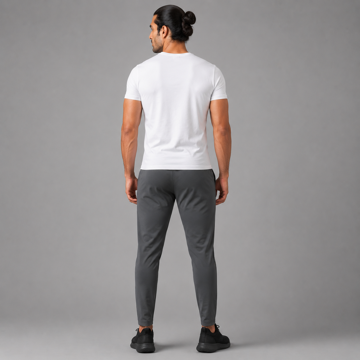 A man viewed from behind wearing Charcoal Grey Trousers, a white short-sleeve t-shirt, dark gray slim-fit pants, and black sneakers, standing against a solid gray studio background.
