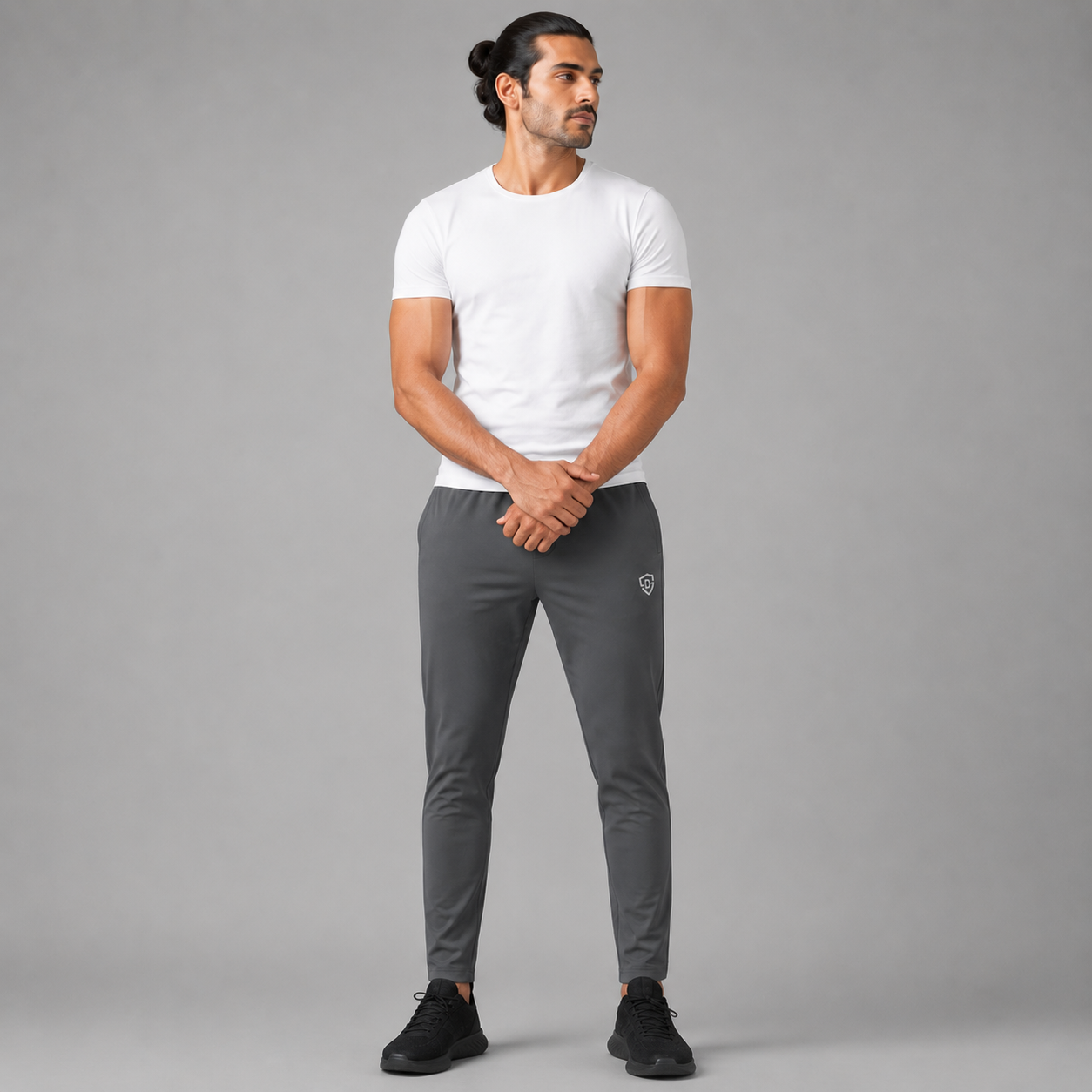 A man modeling a charcoal grey trousers, white crew-neck t-shirt and dark gray athletic jogger pants with black sneakers, standing against a solid gray studio background.